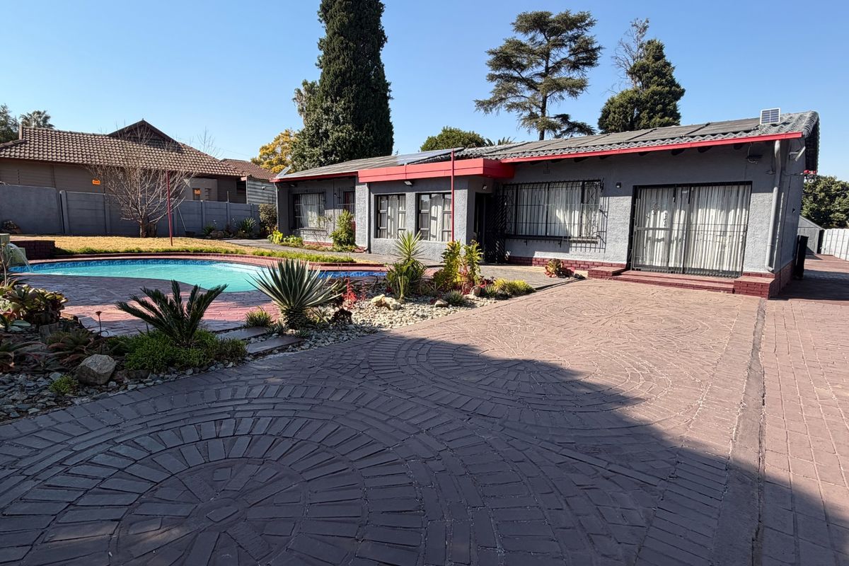 Rear exterior view of the property featuring a paved courtyard, swimming pool, and a single-story building with security bars and solar panels.