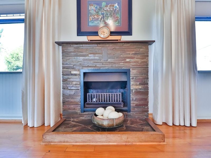 A cozy living room featuring a prominent stone fireplace with a timber mantelpiece and polished timber flooring.