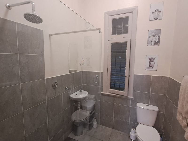 A compact bathroom featuring grey wall and floor tiles, a glass shower screen, a wall-mounted basin, and a standard toilet.