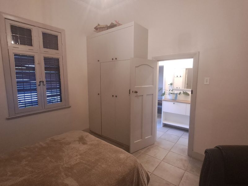 A bedroom featuring a built-in wardrobe, tiled flooring, and a timber-framed window, with an adjoining view into a bathroom area.