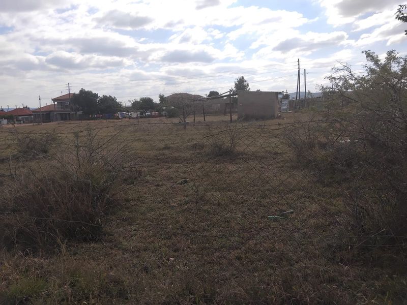 A vacant residential plot featuring overgrown grass, wire mesh perimeter fencing, and a small existing outbuilding structure.