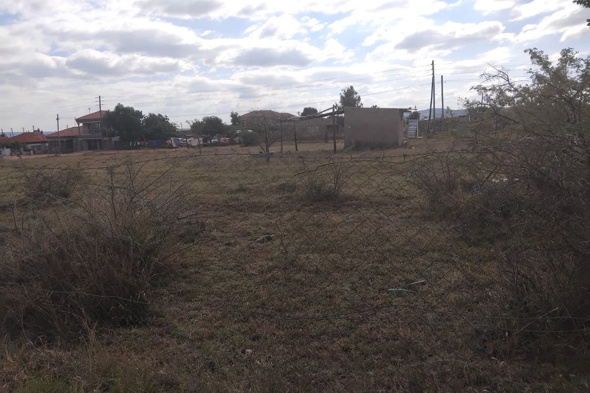 A vacant residential plot featuring overgrown grass, wire mesh perimeter fencing, and a small existing outbuilding structure.