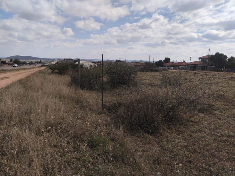 A view of a vacant residential plot featuring natural grass, overgrown shrubs, and perimeter wire fencing, situated near a local road.