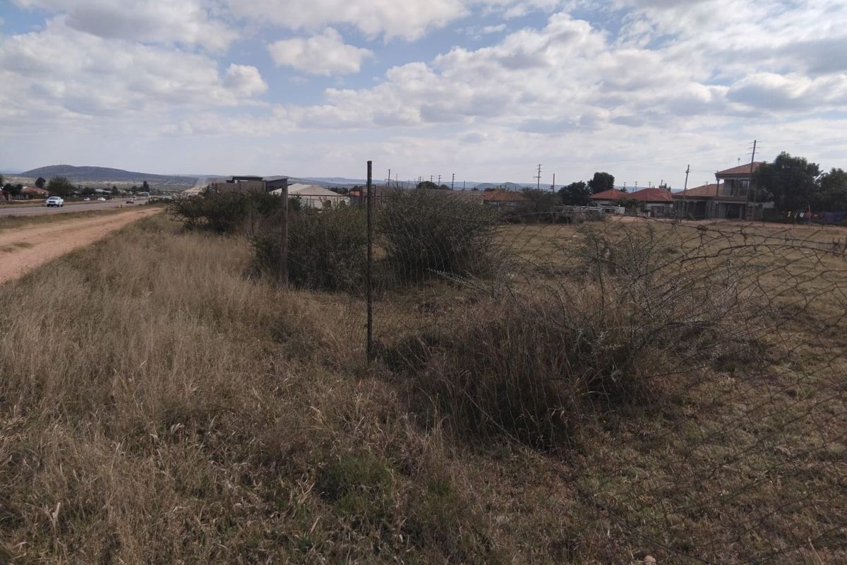 A view of a vacant residential plot featuring natural grass, overgrown shrubs, and perimeter wire fencing, situated near a local road.
