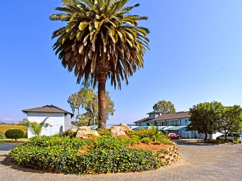 A well-maintained apartment complex entrance featuring a paved driveway, landscaped garden with a central palm tree, and covered parking areas.