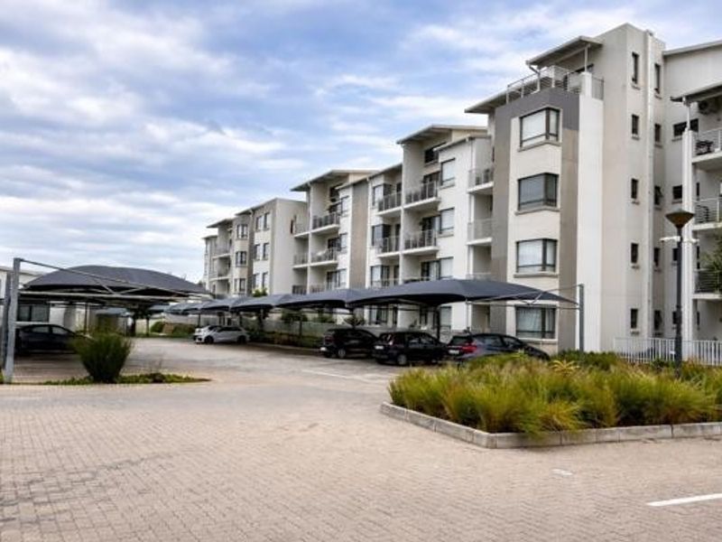 Exterior view of a modern multi-story apartment complex featuring paved parking areas with shade cloth carports and landscaped garden beds.