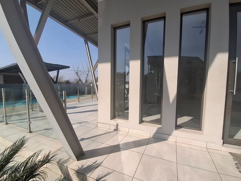 A modern covered patio area featuring tiled flooring, a sleek glass balustrade, and structural steel supports, overlooking a private swimming pool.