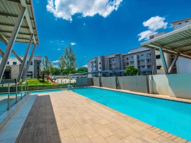 A well-maintained communal swimming pool area featuring paved decking, glass balustrades, and covered seating structures within an apartment complex.