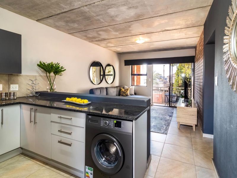 Modern open-plan apartment featuring an exposed concrete ceiling, granite kitchen countertops, integrated laundry, and an exposed brick wall leading to a private balcony.