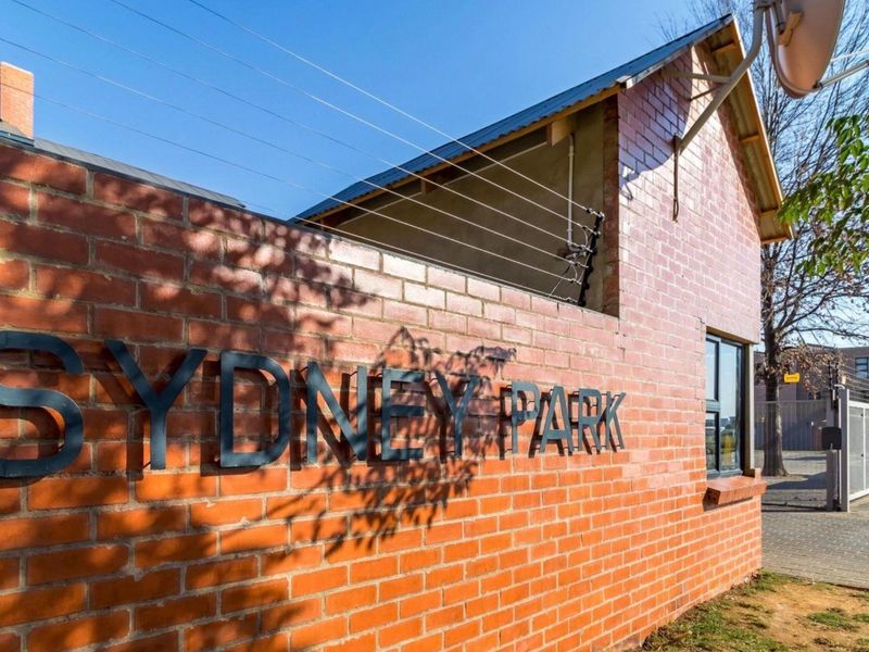 Exterior view of the Sydney Park apartment complex featuring a classic red brick facade, secure electric fencing, and a paved entrance area.