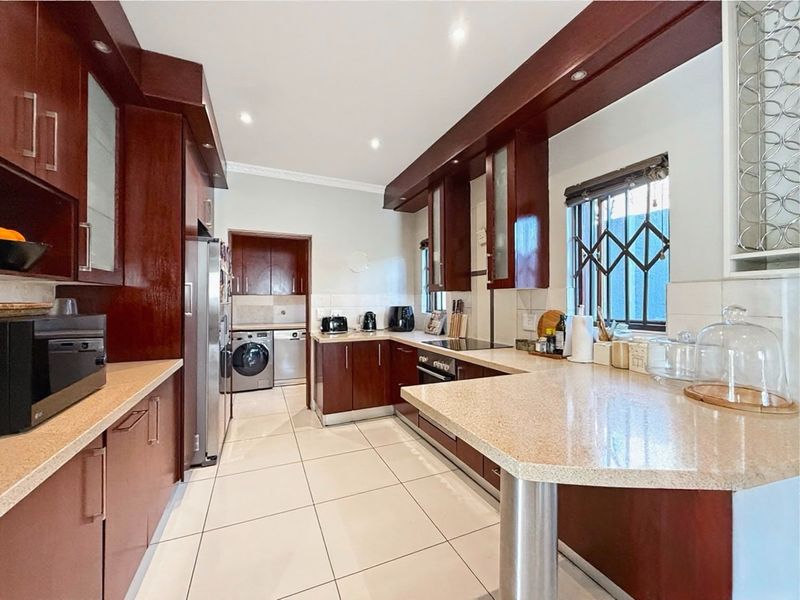 A well-maintained kitchen featuring dark wood cabinetry, stone countertops, integrated appliances, and a functional layout with an adjoining scullery area.
