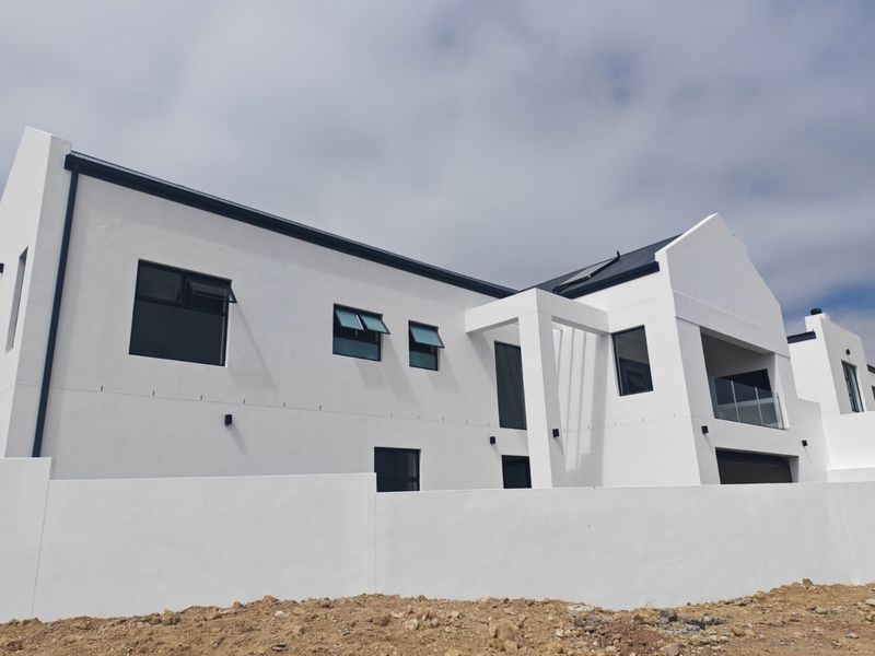 A modern, newly constructed white house featuring clean architectural lines, solar panels, and a glass balcony, set against a clear sky.