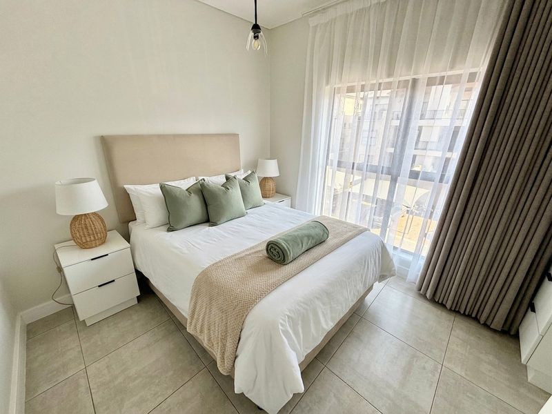 A well-maintained bedroom featuring neutral tiled flooring, a large window with floor-to-ceiling curtains, and modern pendant lighting.