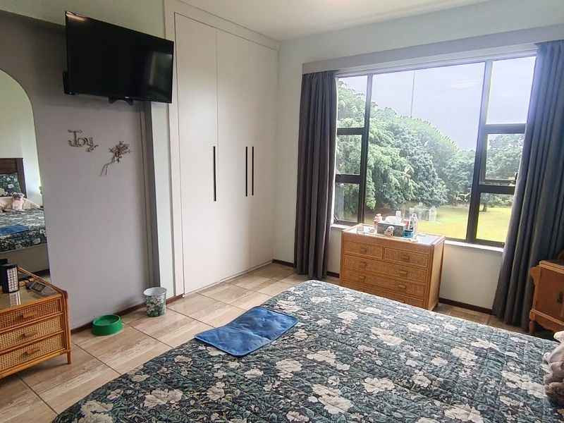 A well-lit bedroom featuring built-in cupboards, tiled flooring, and a large window offering a pleasant garden view.