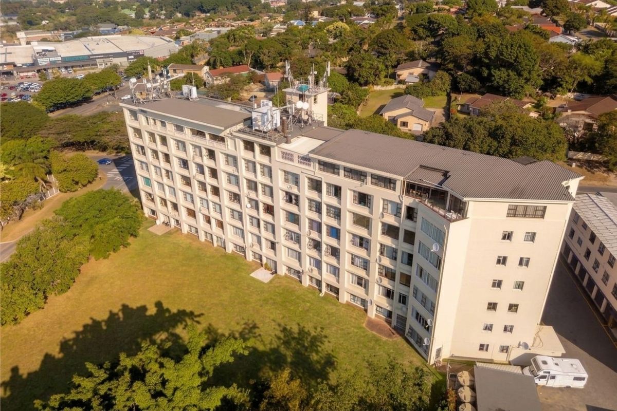 An aerial view of a multi-story apartment building featuring a pitched roof, telecommunication infrastructure, and surrounding communal grounds.