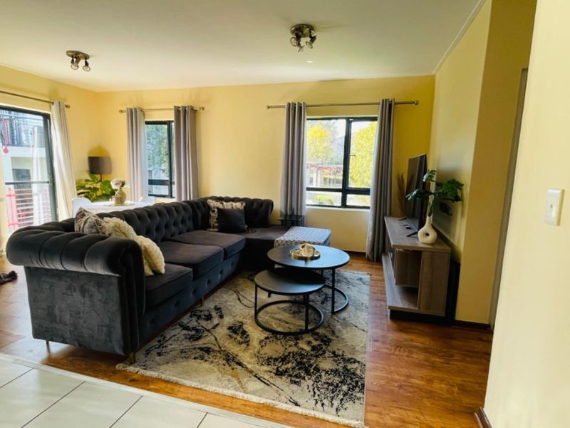 A well-lit living room featuring laminate flooring, neutral walls, and large windows with aluminium frames, providing access to an outdoor area via a sliding door.