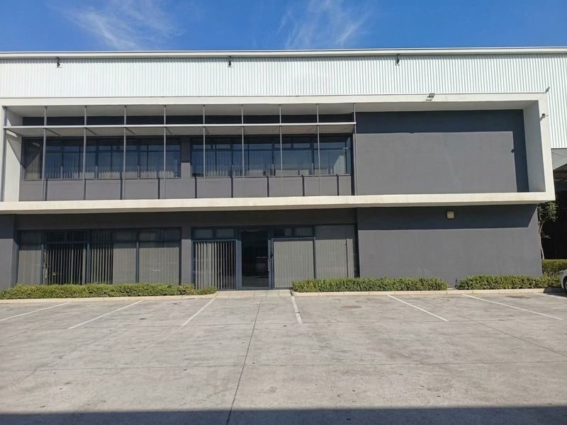 Modern industrial building facade featuring metal cladding, large aluminum-framed windows with sun shading, and a paved concrete parking area.