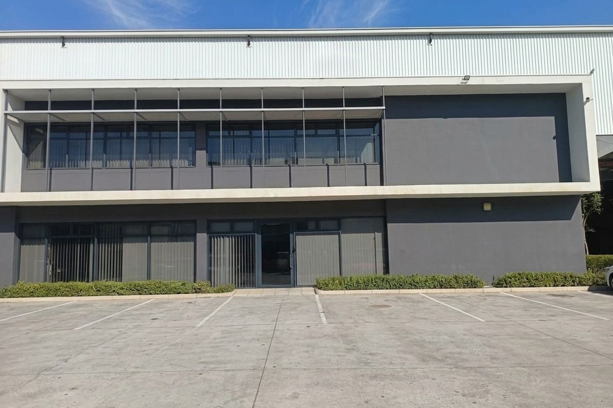 Modern industrial building facade featuring metal cladding, large aluminum-framed windows with sun shading, and a paved concrete parking area.