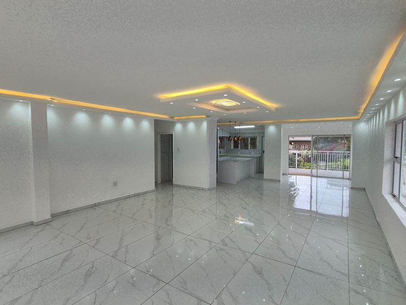 Spacious open-plan living and kitchen area featuring modern recessed cove lighting, polished marble-look porcelain floor tiles, and seamless access to an outdoor balcony.