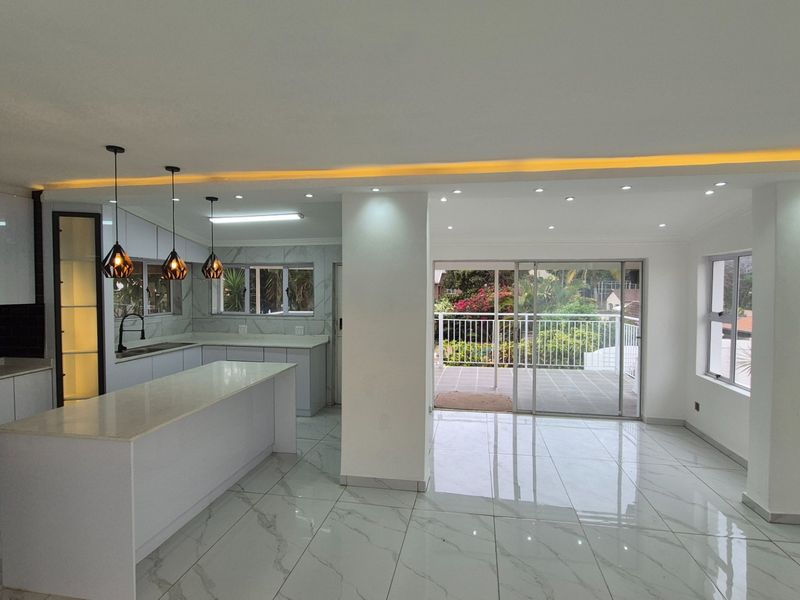 Modern open-plan kitchen and living area featuring sleek white cabinetry, stone countertops, recessed lighting, and large sliding doors leading to an outdoor patio.