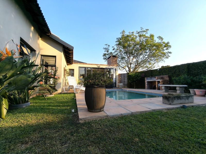 A well-maintained outdoor pool area featuring a paved deck, built-in brick braai, and established garden surroundings, perfect for outdoor entertaining.