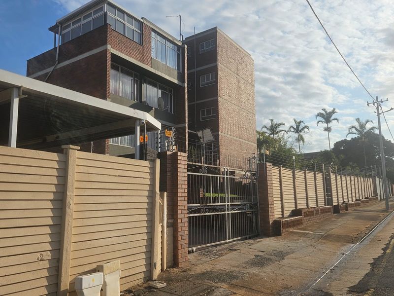 Exterior view of a multi-story brick apartment building featuring a secure perimeter wall, electric fencing, and a gated entrance.