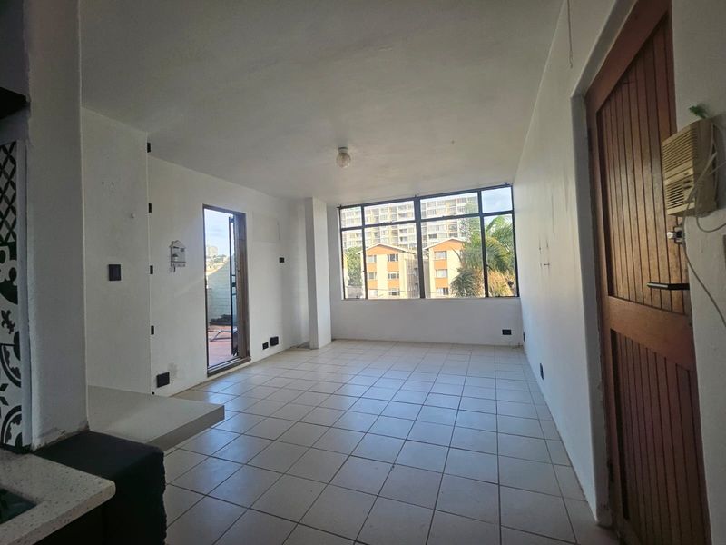 An empty open-plan living area featuring tiled flooring, large windows providing natural light, and direct access to an outdoor balcony space.