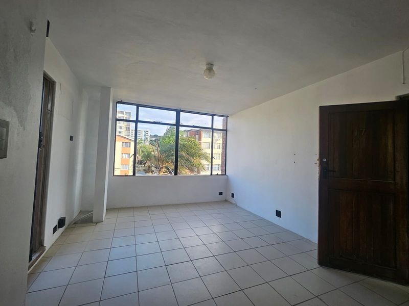 An empty, sunlit living area featuring tiled flooring, a large window with neighborhood views, and a traditional timber entrance door.