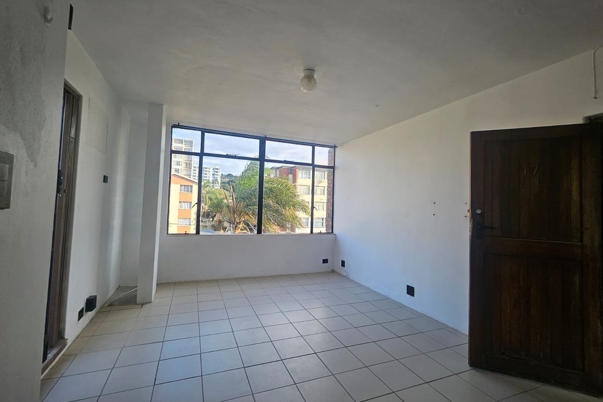 An empty, sunlit living area featuring tiled flooring, a large window with neighborhood views, and a traditional timber entrance door.