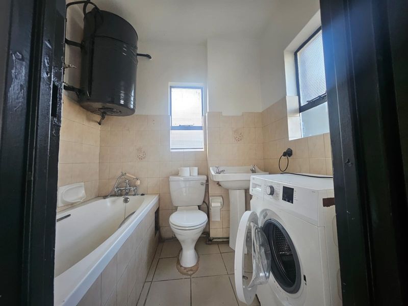 A functional bathroom featuring a bathtub, pedestal basin, and toilet, with wall-mounted geyser and tiled finishes. The space requires cosmetic updates.