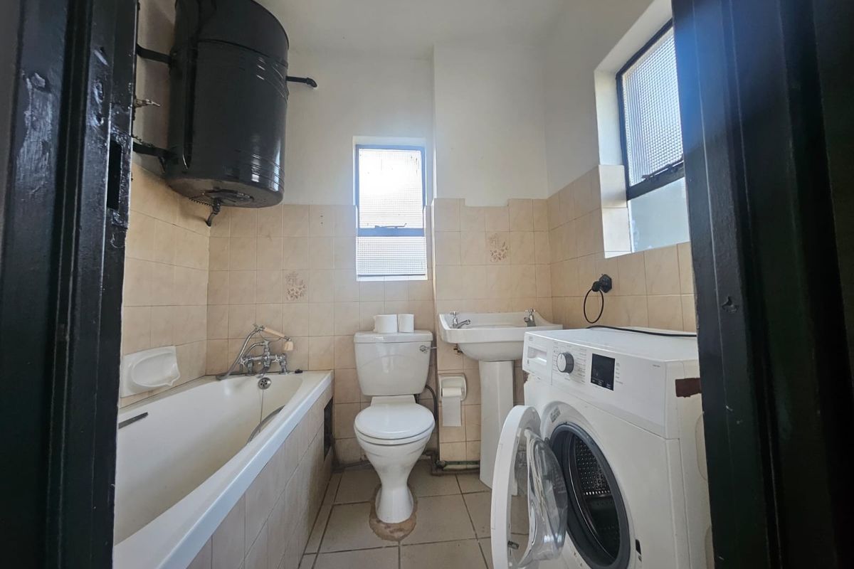 A functional bathroom featuring a bathtub, pedestal basin, and toilet, with wall-mounted geyser and tiled finishes. The space requires cosmetic updates.