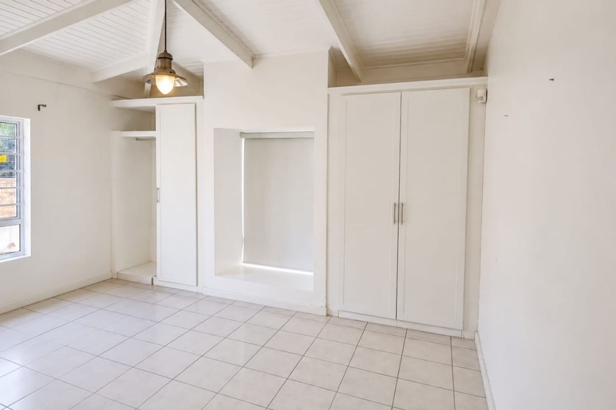 A bright, empty bedroom featuring tiled floors, exposed beam ceilings, and ample built-in cupboard storage with security bars on the window.
