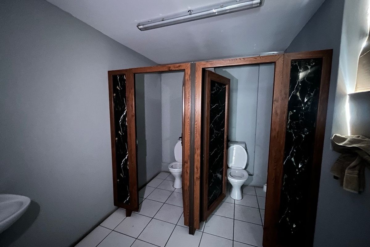Industrial bathroom facility featuring two toilet cubicles with wooden frames and marble-effect panels, tiled flooring, and a wall-mounted washbasin.