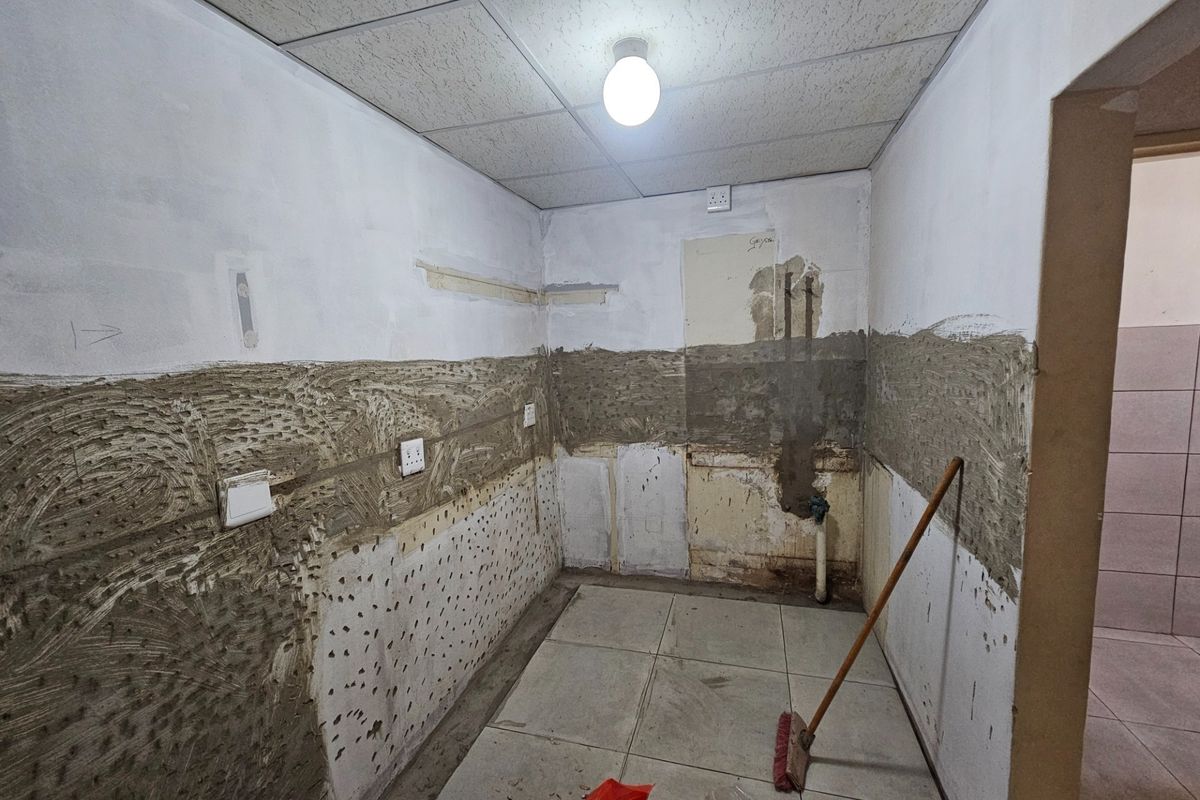 An empty kitchen space currently undergoing renovation, featuring exposed wall substrates, tiled flooring, and a suspended ceiling with basic lighting.