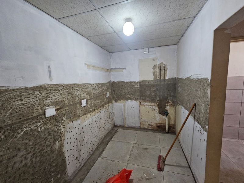 An empty kitchen space currently undergoing renovation, featuring exposed wall substrates, tiled flooring, and a suspended ceiling with basic lighting.