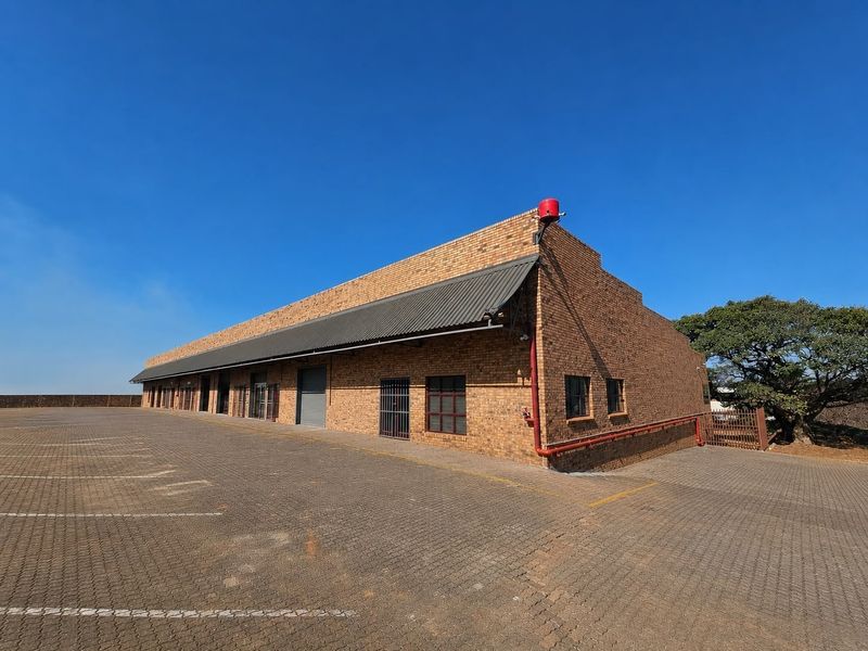 A commercial warehouse building featuring a face brick facade, corrugated metal roofing, roller shutter access, and an extensive paved parking area.