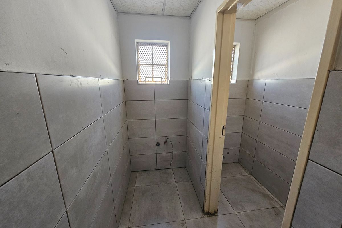 An empty commercial bathroom space featuring tiled walls and floors, security window grilles, and exposed plumbing connections ready for fixture installation.