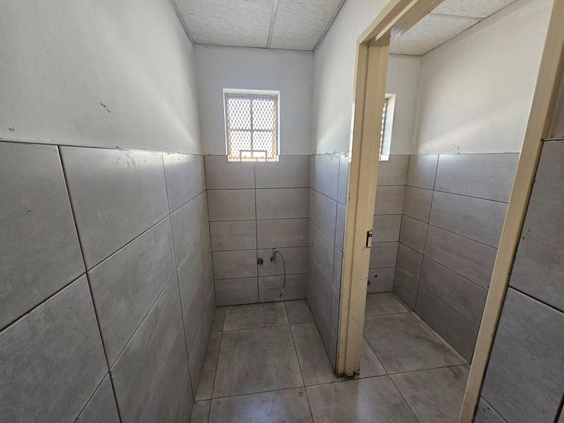 An empty commercial bathroom space featuring tiled walls and floors, security window grilles, and exposed plumbing connections ready for fixture installation.