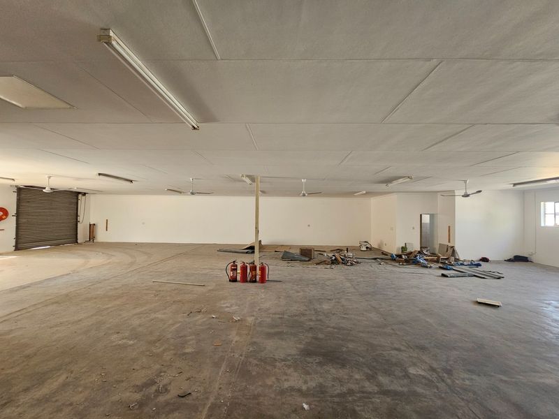 Spacious open-plan commercial utility area featuring a roller shutter door, ceiling fans, and fluorescent lighting, suitable for storage or workshop use.