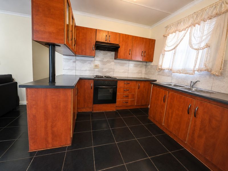 A functional kitchen featuring wood-finish cabinetry, dark countertops, built-in oven with gas hob, extractor fan, and dark tiled flooring.
