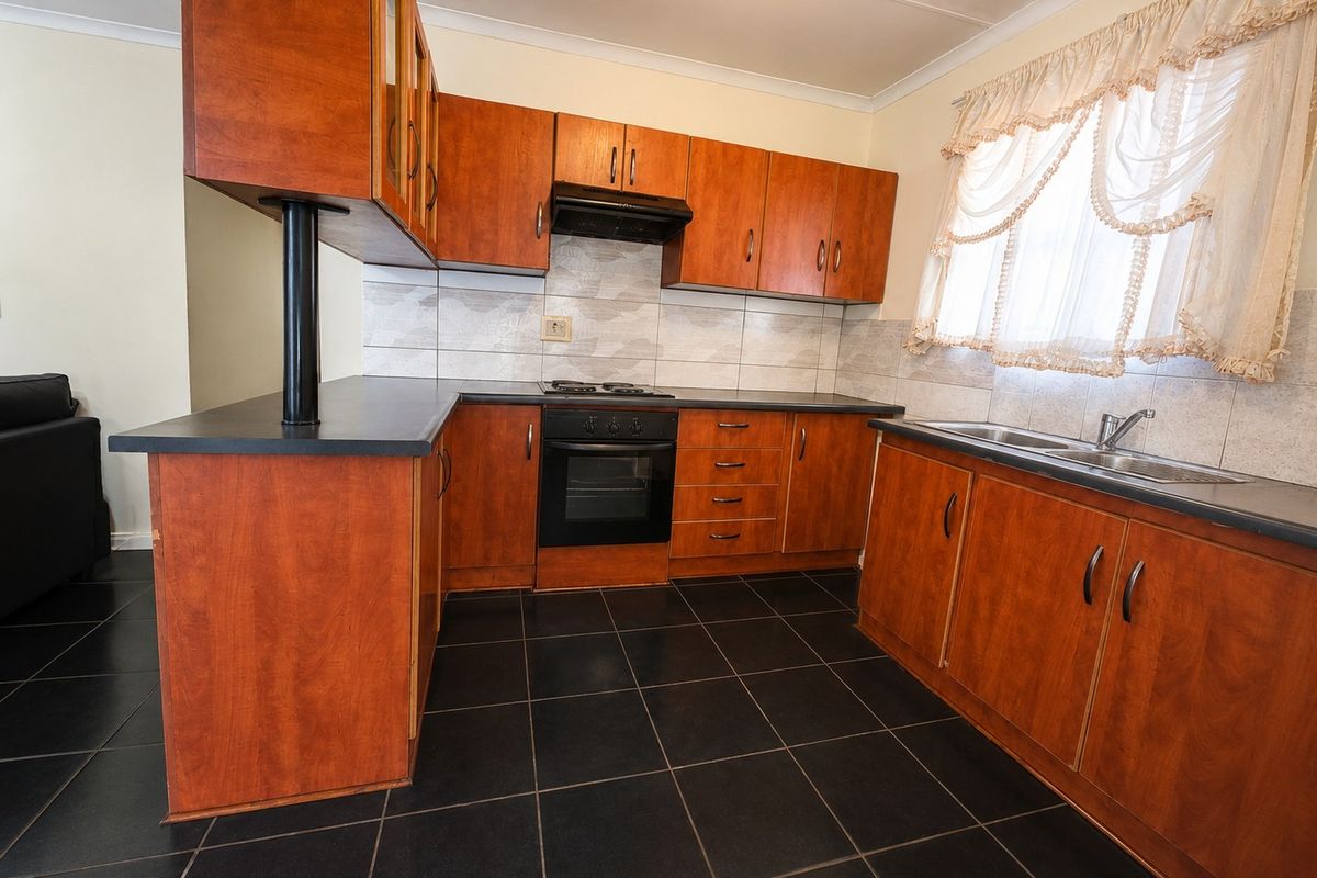 A functional kitchen featuring wood-finish cabinetry, dark countertops, built-in oven with gas hob, extractor fan, and dark tiled flooring.