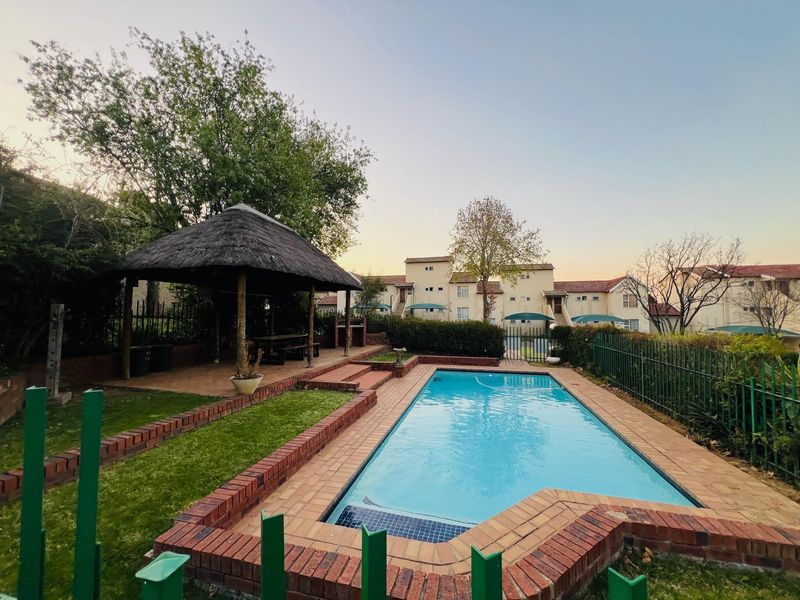 Communal pool area featuring a swimming pool, brick paving, and a thatch lapa with a built-in braai, set within a residential apartment complex.