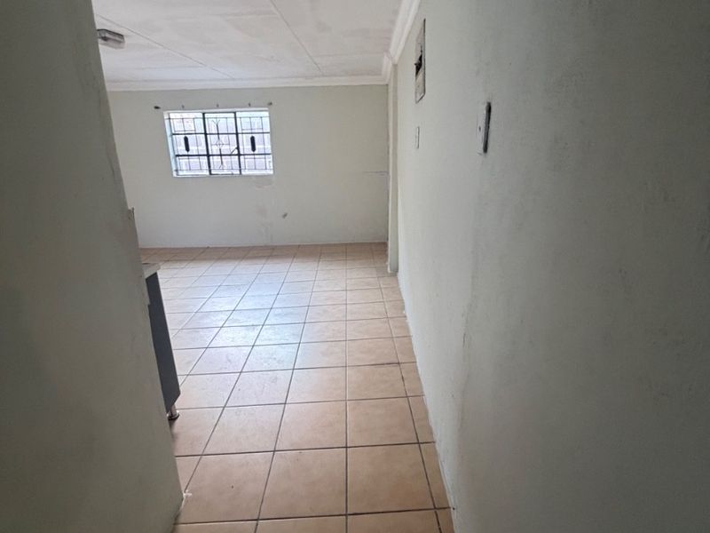 An empty open-plan living area featuring tiled flooring, coved ceilings, and a window with security bars, requiring cosmetic updates to improve its appeal.