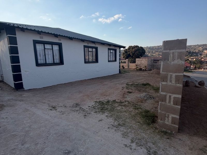 Exterior view of a single-story house featuring a corrugated metal roof, aluminium window frames, and an unpaved yard with an unfinished masonry pillar.