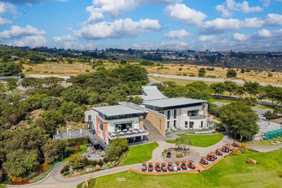 Aerial view of a modern multi-level clubhouse facility featuring stone cladding, metal roofing, and extensive landscaped grounds with paved access paths.