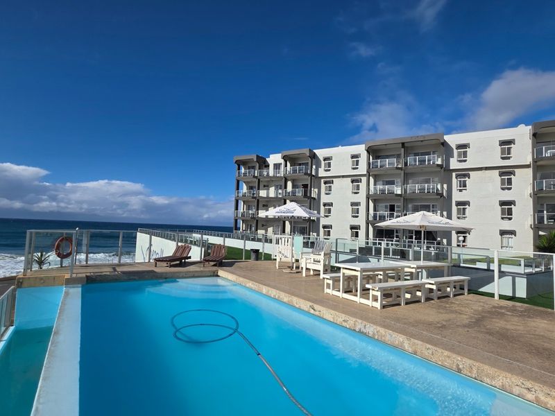 Outdoor pool area featuring a swimming pool with glass balustrades, overlooking the ocean and adjacent to the apartment building complex.