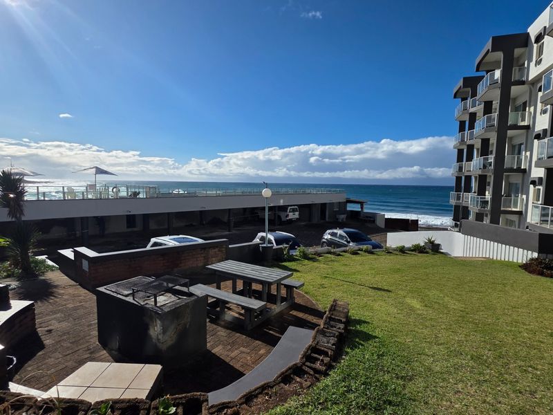 A well-maintained rear exterior area featuring a paved patio with a built-in braai, landscaped lawn, and direct views of the ocean and apartment complex.