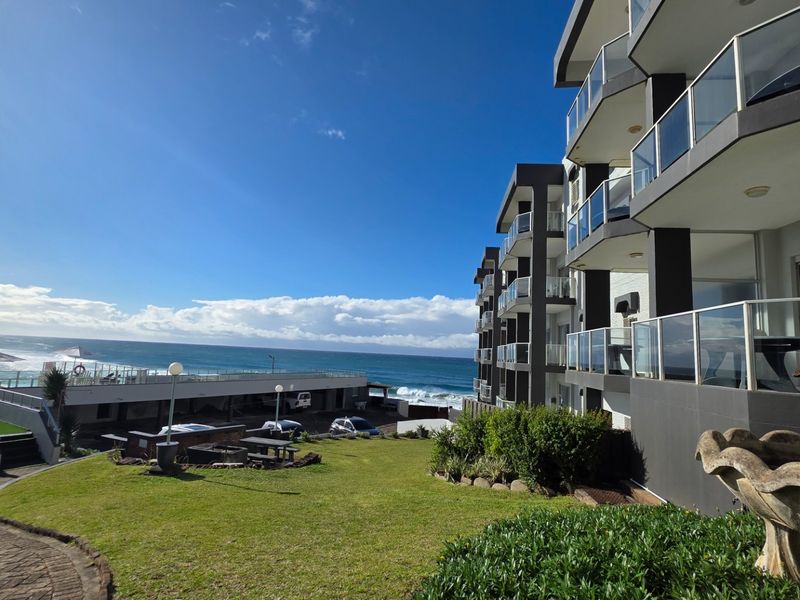 Rear view of the apartment complex featuring tiered balconies with glass balustrades, overlooking a landscaped garden and the ocean.