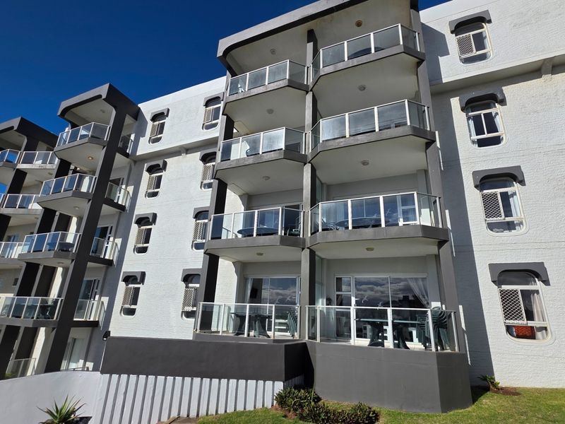 A multi-story apartment building exterior featuring stacked balconies with glass balustrades and sliding door access, set against a clear blue sky.