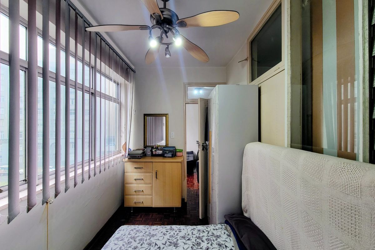 A compact bedroom featuring parquet flooring, a ceiling fan, and large windows with vertical blinds, offering a functional space with potential for modernization.